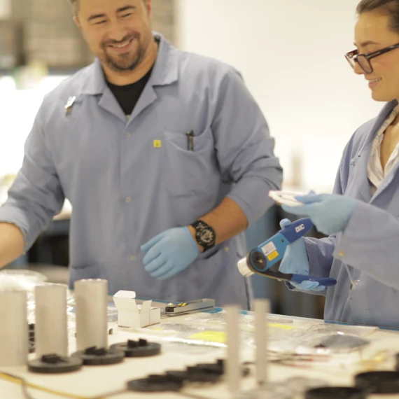 A male employee dressed in a blue smock and blue gloves and a female employee dressed in a blue smock and blue gloves assembling antennas.