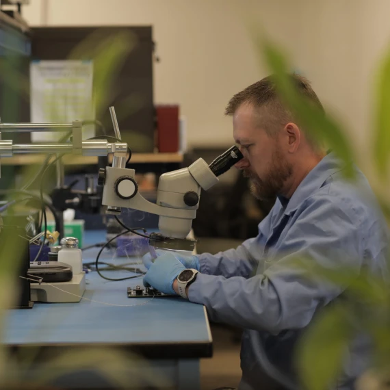A male technician dressed in a blue smock with blue gloves is seen soldering a board in the background with blurred plants in the foreground. The male employee is looking into a microscope, analyzing the board.